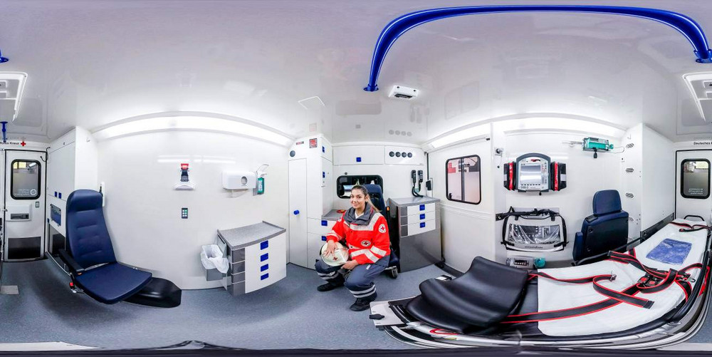 Auf dem Foto ist eine 3D-Aufnahme des Inneren eines Rettungswagens zu sehen, in dem eine Person in Einsatzkleidung hockt und lächelt.