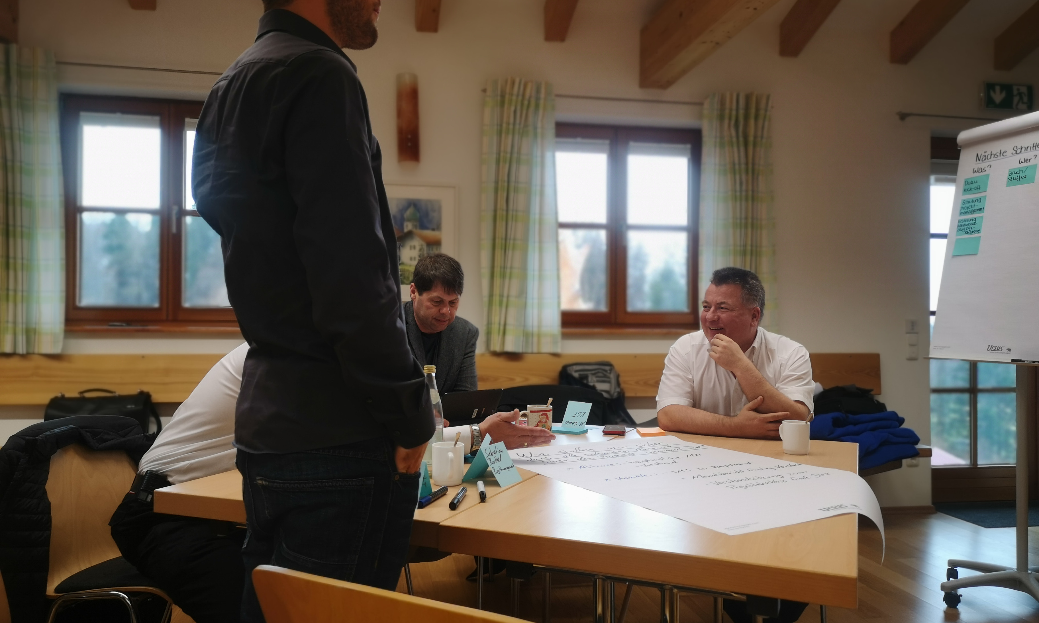Kick-off-Workshop_KV-Rosenheim_02-2020_von_Susanne-Bruch_DRK Drei Menschen sitzen um einen Tisch, davor steht eine weitere. Auf dem Tisch sind Marker, Flipchart-Papier und weitere Büromaterialien.