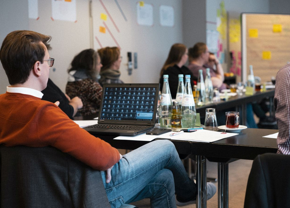 Foto aus einem Seminarraum. Ein Mann sitzt in einer Runde vor seinem Laptop.