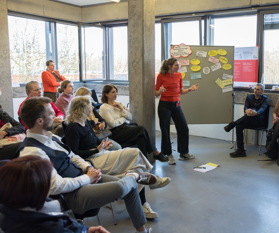 Frau steht vor einer Gruppe sitzender Personen in einem hellen Raum mit großen Fenstern und einem Flipchart mit bunten Notizzetteln