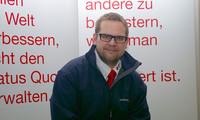 Mann mit dunkelblauer Jacke und roter Krawatte vor weißer Wand mit rotem Text.