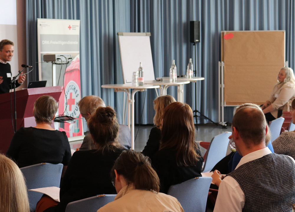 Person steht an einem Rednerpult vor Publikum in einem Konferenzraum mit Flipcharts und Rollup im Hintergrund