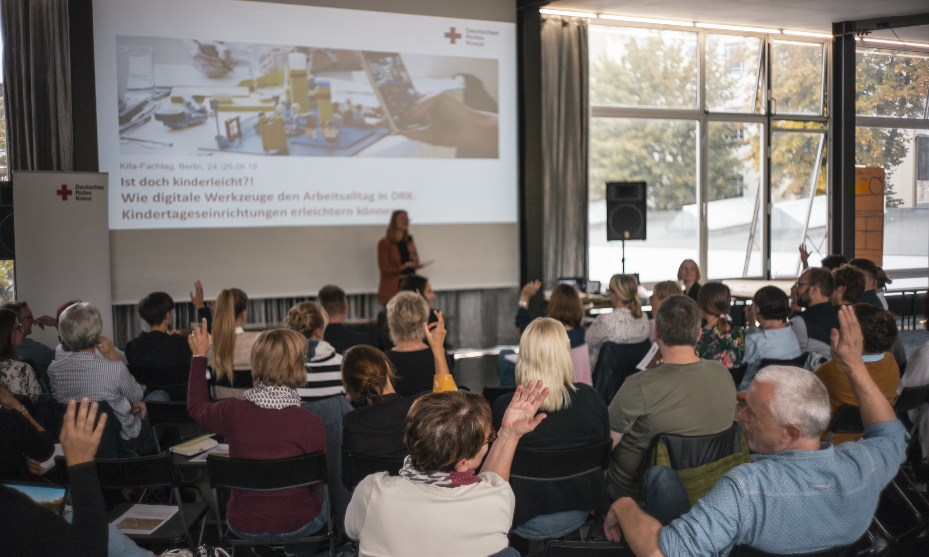 Frieder Unselt_DRK_Fachtagung_Kita_Publikum-Präsentation Foto der Kita-Fachkonferenz. Gruppe von Teilnehmenden meldet sich und vor der Gruppe wird eine Präsentation druchgeführt.