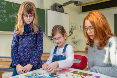 Zwei Mädchen sitzen und stehen mit einer erwachsenen Frau an einem Tisch und beschäftigen sich gemeinsam mit einem Lernspiel. Im Hintergrund ist ein Klassenraum zu sehen.