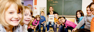 Auf dem Foto ist eine Schulsituation abgebildet. Kinder sitzen mit ihrer Lehrerin in einem Stuhlkreis und die Lehrerin hält eine Schild hoch, auf dem steht: "Kinder helfen Kindern.".
