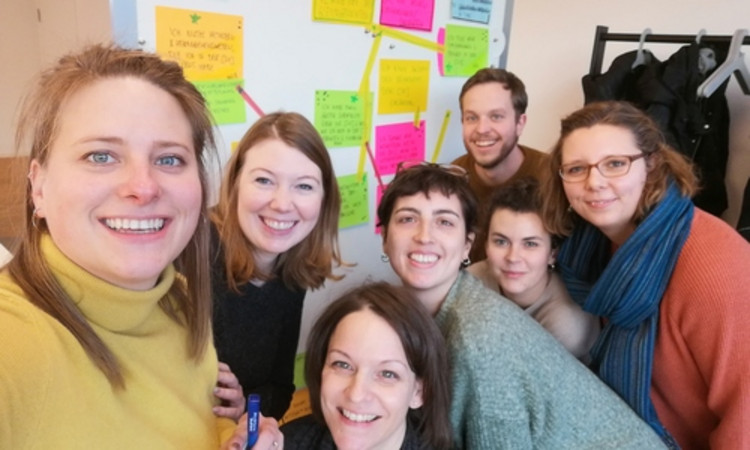 Sieben Menschen posieren vor einer weißen Wand mit bunten Post-Its für ein Selfie und lächeln.