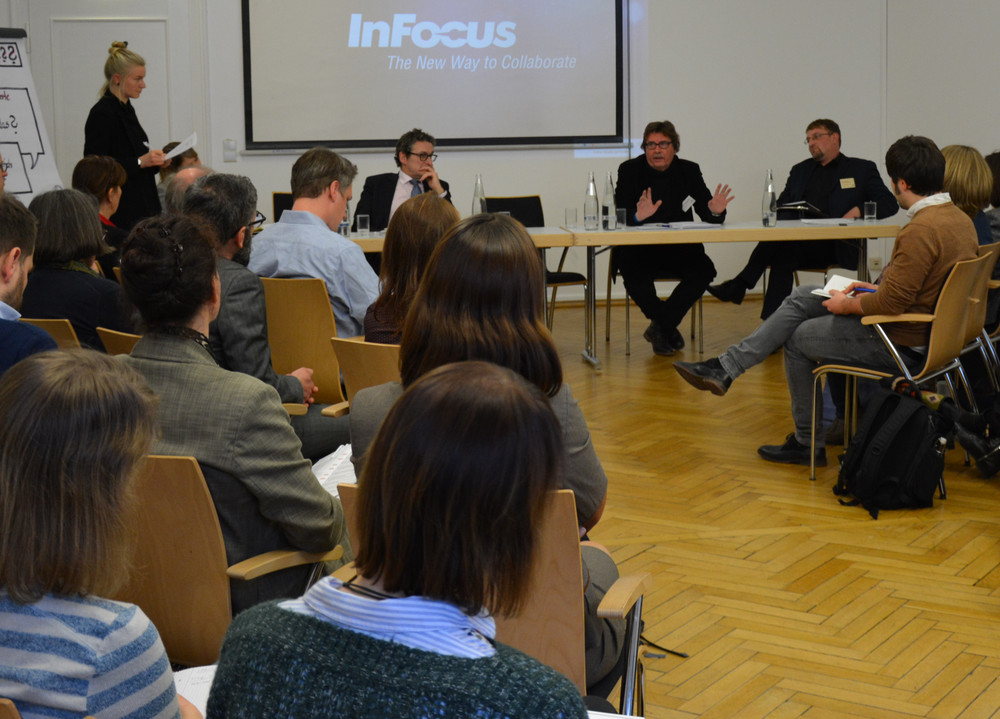 Mehrere Personen sitzen in einem Raum mit Holzboden und hören einer stehenden Frau zu, die vor einem Flipchart spricht.