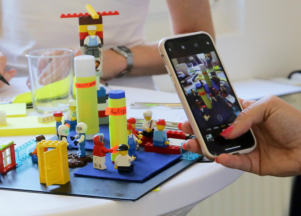 Zwei Personen stehen an einem Stehtisch. eine fotografiert die mit Lego inszenierte Baustelle