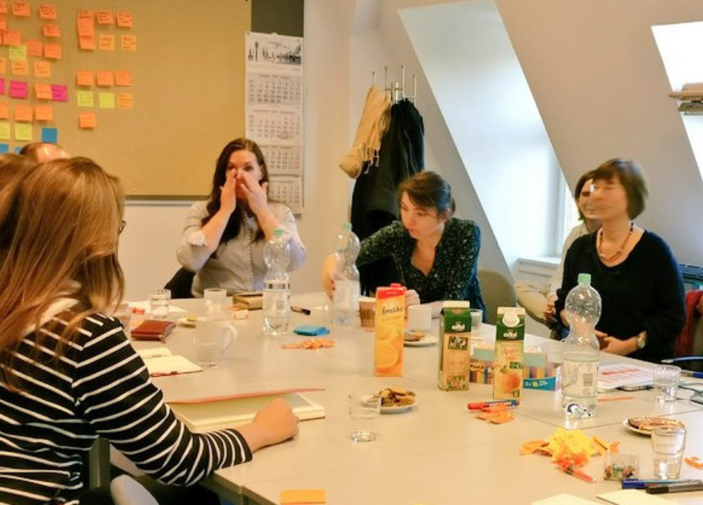 Mehrere Personen sitzen an einem Tisch mit Getränken und Notizzetteln, ein Mann steht und gestikuliert neben einem Laptop.