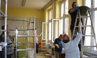 Mehrere Personen renovieren mit Leitern und Farbeimern einen Raum mit hohen Fenstern, 