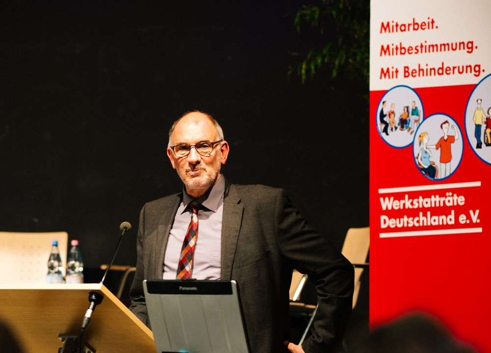 Mann im Anzug steht an Rednerpult mit Mikrofon und Laptop, im Hintergrund Banner von Werkstatträte Deutschland e.V.