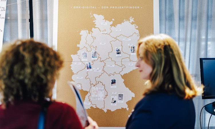 Man sieht eine Korkwand mit einer Deutschlandkarte und dem Titel "DRK-Digital - Der Projektfinder" und verschiedenen angepinnten Kärtchen vor dem zwei Frauen im Gespräch stehen.
