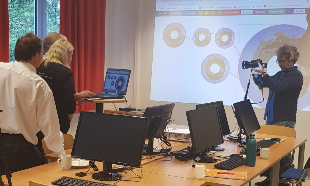 Im Raum stehe mehrere Computer, vor einem Projektorbild fotografiert eine Person mehrere Personen an einem Stehpult.