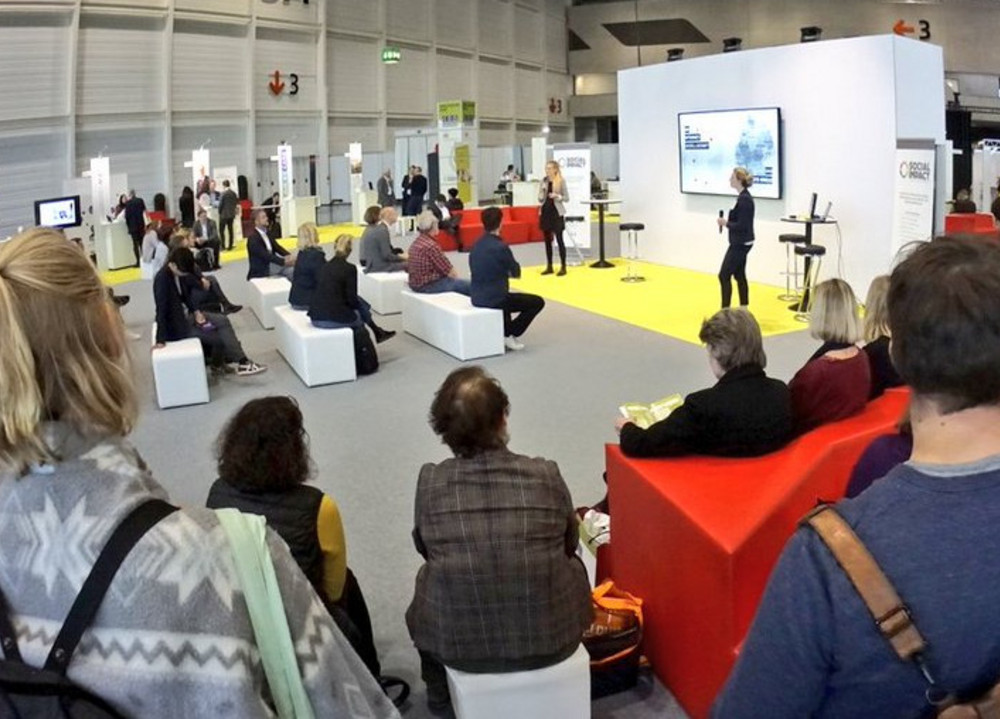 Menschen sitzen und stehen in einer Halle vor einer Bühne mit Präsentation und großem Bildschirm