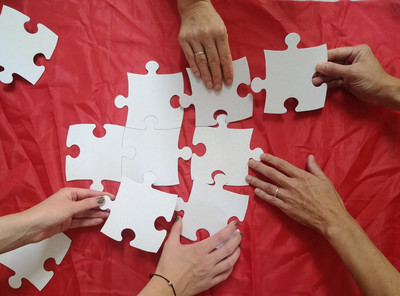 Es liegen weiße Puzzleteile auf einem roten Tuch aus, die von mehreren Händen aufgegriffen und zusammengesteckt werden.