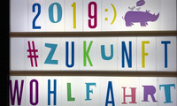 Leuchtkasten mit bunten Buchstaben und Zahlen, die "2019 :) #ZUKUNFT WOHLFAHRT" bilden, dazu eine lila Nilpferd-Illustration mit zwei Vögeln und Sprechblase.