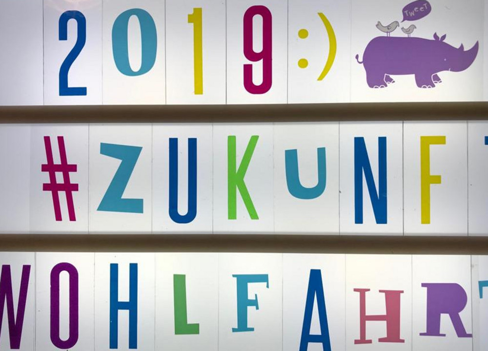 Leuchtkasten mit bunten Buchstaben und Zahlen, die "2019 :) #ZUKUNFT WOHLFAHRT" bilden, dazu eine lila Nilpferd-Illustration mit zwei Vögeln und Sprechblase.