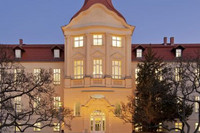 Beleuchtetes Gebäude des DRK Generalsekretariats mit Turm und roten Dachziegeln, umgeben von Bäumen und einem Weg mit Bänken im Vordergrund.