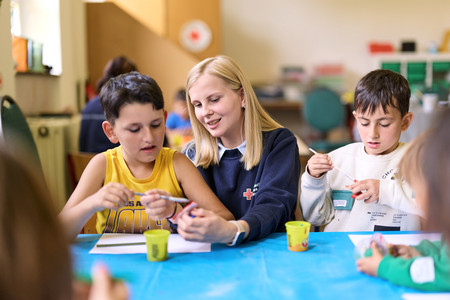 Zwei Kinder und eine junge Freiwillige sitzen an einem Tisch mit blauer Tischdecke und malen mit Pinseln und gelben Bechern Farbe.