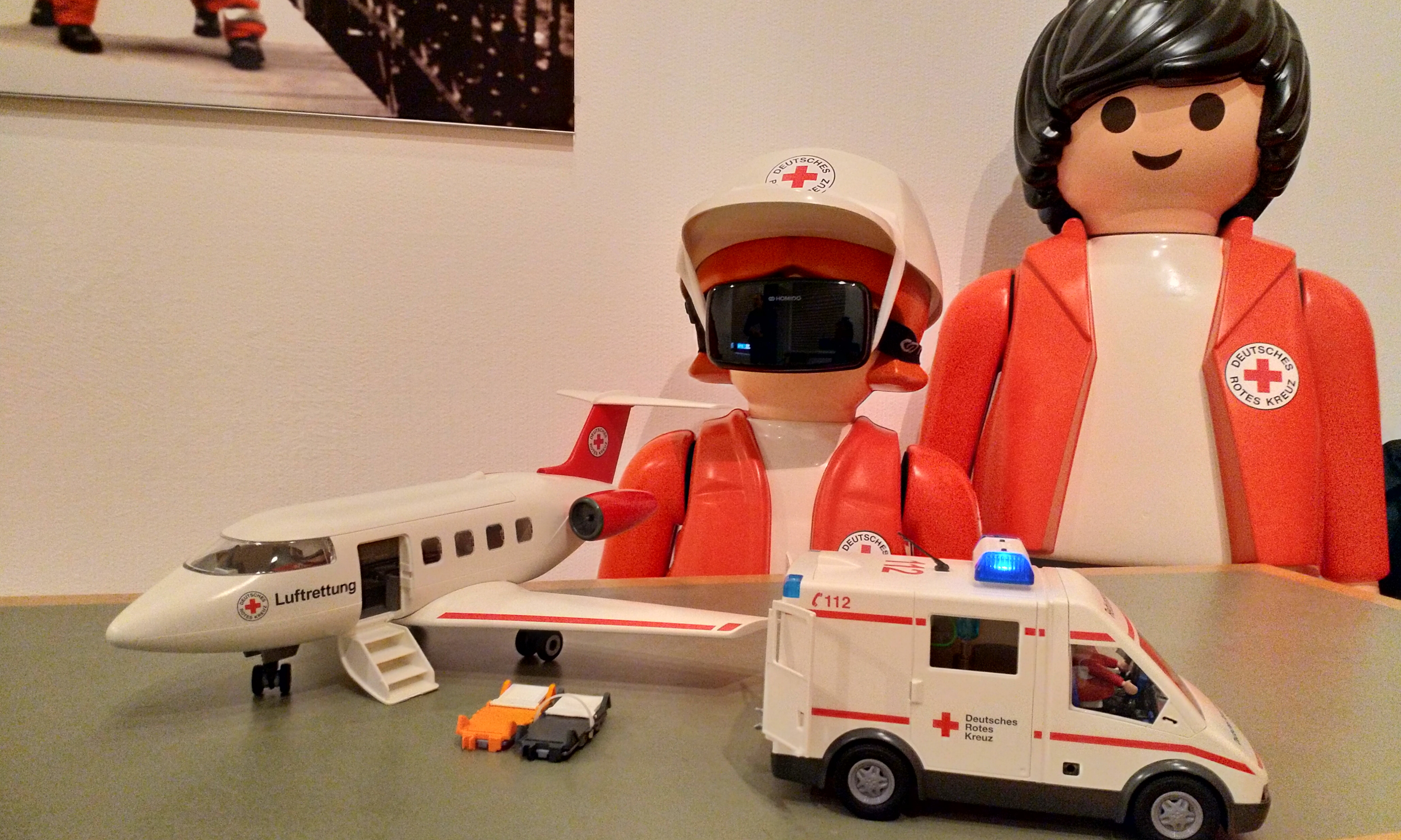Zwei größe Playmobil-DRK-Figuren stehn vor einem kleinem Playmobil-DRK-Flugzeug und DRK-Rettungswagen.