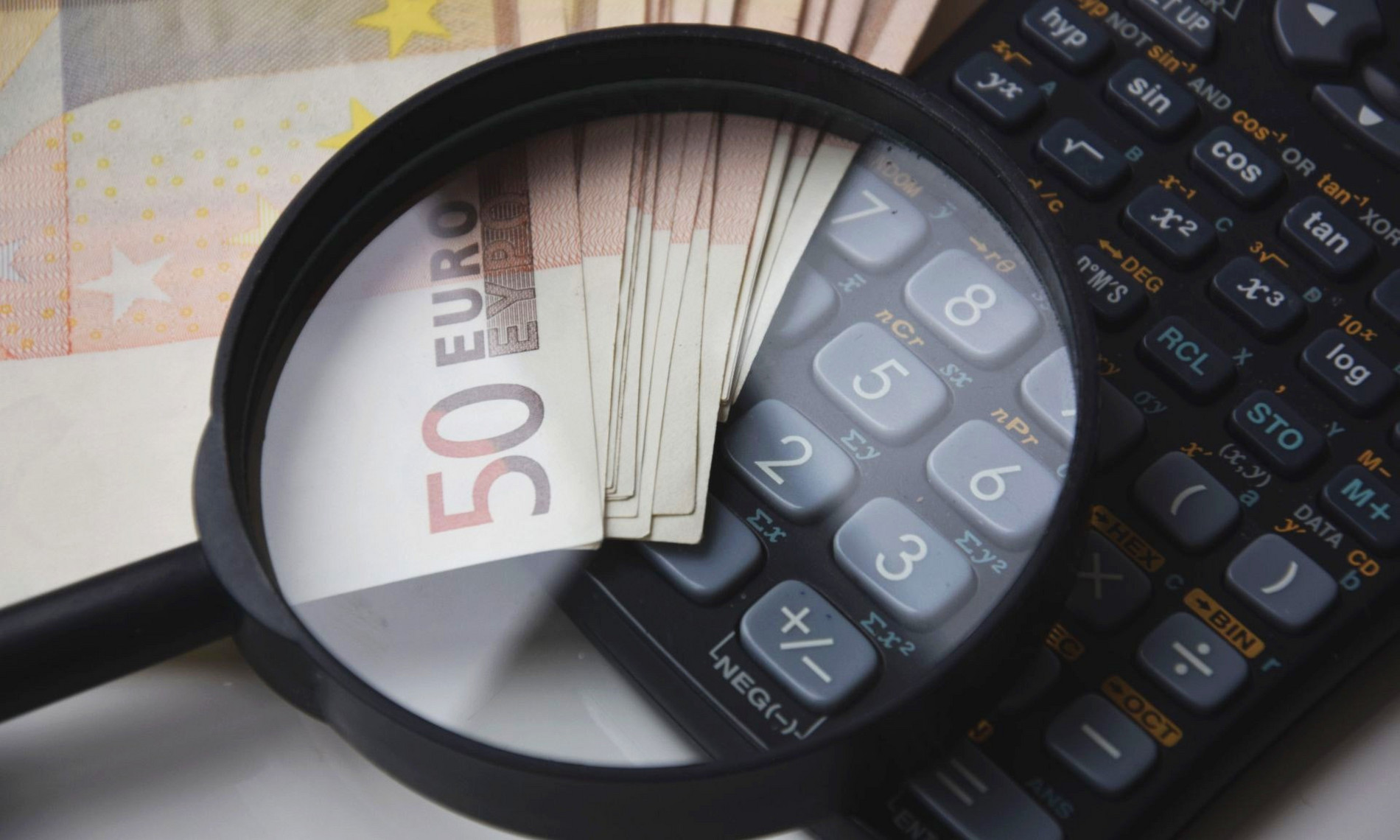 Eine Lupe bildet ein Bündel 50 Euro-Scheine und einen Taschenrechner ab. So entsteht eine Assoziation mit den Themen Finanzen, Überschuldung und Schuldnerberatung.