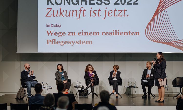 Auf dem Bild sitzen 5 Personen auf einem Podium, eine Person steht. Im Hintergrund sieht man eine Präsentation mit dem Titel "Im Dialog: Wege zu einem resilienten Pflegesystem"