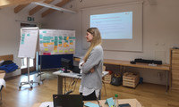 In der Mitte steht Frau Stuffer, im Hintergrund sind Flipchats und ein Beamer-Bild aufgestellt.