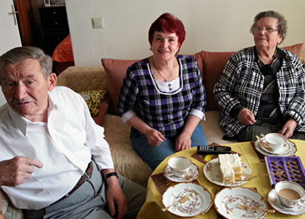 Drei ältere Menschen sitzen auf einem Sofa und an einem Tisch mit Tassen, Tellern, Kuchen und Pralinen in einem Wohnzimmer.