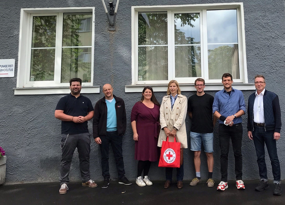 Gruppe aus sieben Personen steht vor einem Gebäude mit Rotem Kreuz