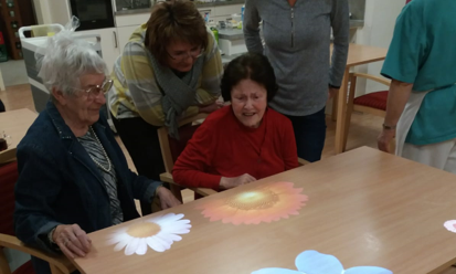 Einsatz der Tovertafel im BRK Seniorenhaus Kronach. Zwei ältere Menschen sitzen an einem Tisch, auf dem Blumen digital projiziert werden.
