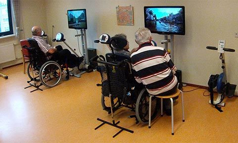 In einem Raum stehen zwei Heimtrainer verbunden mit Bildschirmen (Bike-Labs) an denen Seniorinnen und Senioren sitzen.