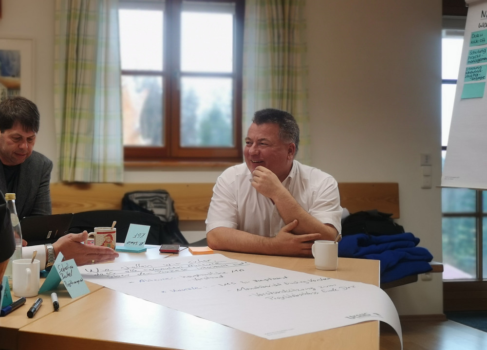 Ein Mitarbeitender des Kreisverband Rosenheim sitzt an einem Tisch vor einem Flipchart und lacht.