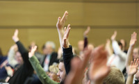 Mehrere erhobene Hände in einem Versammlungsraum mit Holzwand im Hintergrund.
