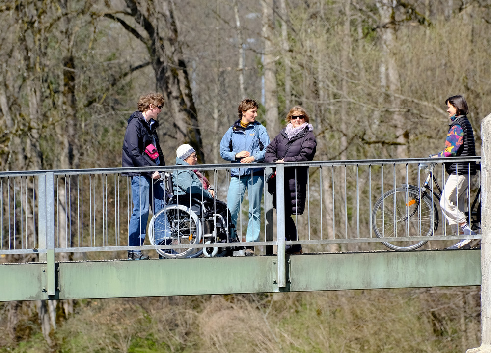 Menschen stehen und gehen auf einer Fußgängerbrücke über einen Fluss, eine Person sitzt im Rollstuhl, im Hintergrund Bäume und Häuser.
