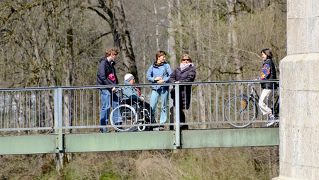 Menschen stehen und gehen auf einer Fußgängerbrücke über einen Fluss, eine Person sitzt im Rollstuhl, im Hintergrund Bäume und Häuser.