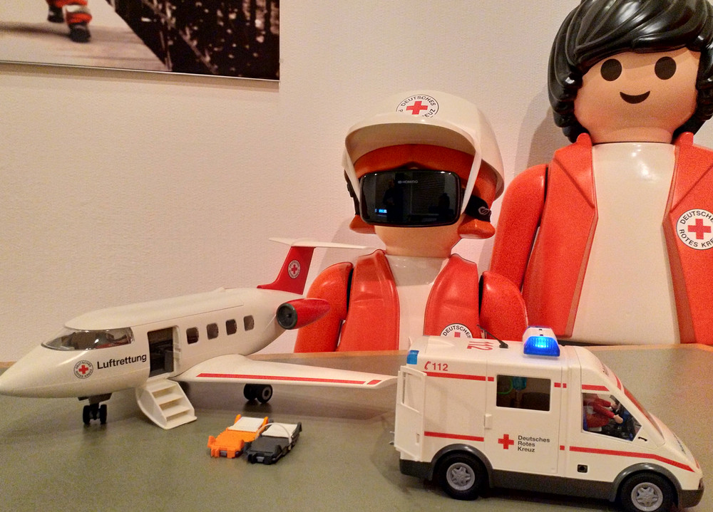 Zwei größe Playmobil-DRK-Figuren stehn vor einem kleinem Playmobil-DRK-Flugzeug und DRK-Rettungswagen.