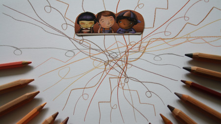 Buntstifte liegen kreisförmig auf weißem Papier. In der Mitte verlaufen zahlreiche Linien in verschiedenen Farben, die von den Stiften ausgehen und sich kreuzen. Am oberen Rand sind zwei gezeichnete Kinderfiguren mit unterschiedlichen Hauttönen und Frisuren zu sehen.