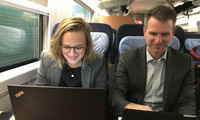 Zwei Personen sitzen nebeneinander in einem Zugabteil, beide arbeiten an Laptops auf einem Tisch vor sich.