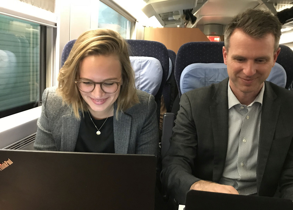 Zwei Personen sitzen nebeneinander in einem Zugabteil, beide arbeiten an Laptops auf einem Tisch vor sich.