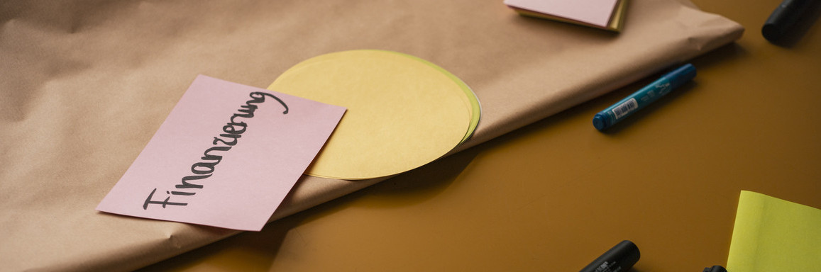 Stifte und Zettel. Ein Zettel in rosa mit der Aufschrift "Finanzierung"