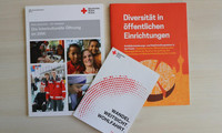Drei Broschüren des Deutschen Roten Kreuzes liegen auf einem hellen Tisch, darunter eine orangefarbene mit dem Titel 'Diversität in öffentlichen Einrichtungen'.
