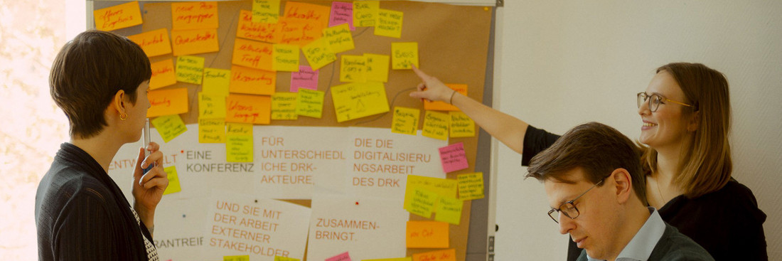 Kompetenzzentren_nehmen_Fahrt_auf Drei Personen stehen vor einem Flipchart mit diversen Post-Its zum Thema Digitalisierung. In der Mitte steht zentral und groß einige Ziele wie das DRK digitalisiert werden könnte.