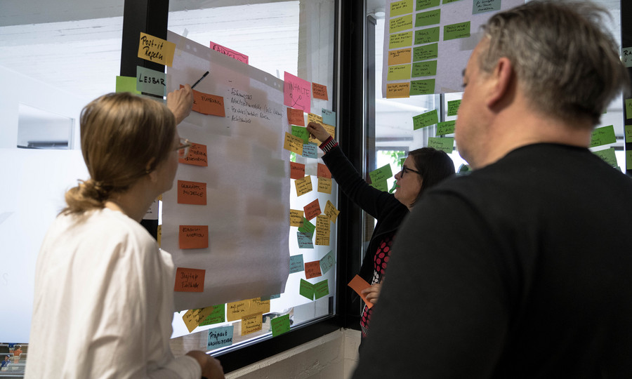 Think und Do Tank in Action Auf dem Foto stehen drei Personen vor einem Fenster mit Flipchart und Post-it.