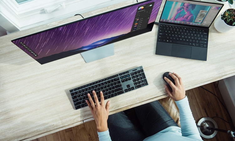 Person sitzt am Schreibtisch und tippt gerade in die Tastatur. Auf dem Schreibtisch ist ein großer Desktop, ein Laptop und eine Computermaus zu sehen.