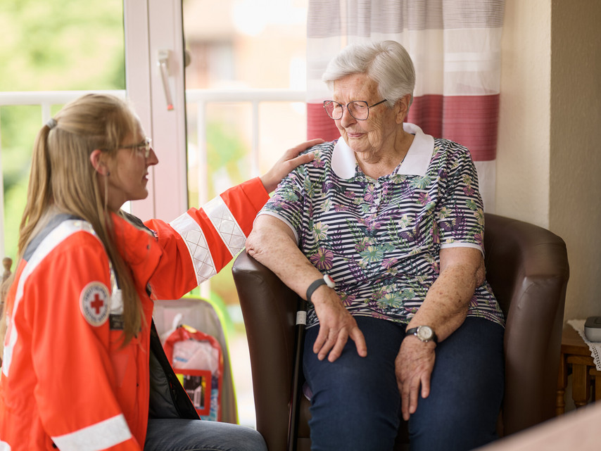 Eine Frau in einer orange-weißen Jacke des Deutschen Roten Kreuzes spricht mit einer älteren Frau, die auf einem Sessel sitzt, und legt ihr tröstend die Hand auf die Schulter