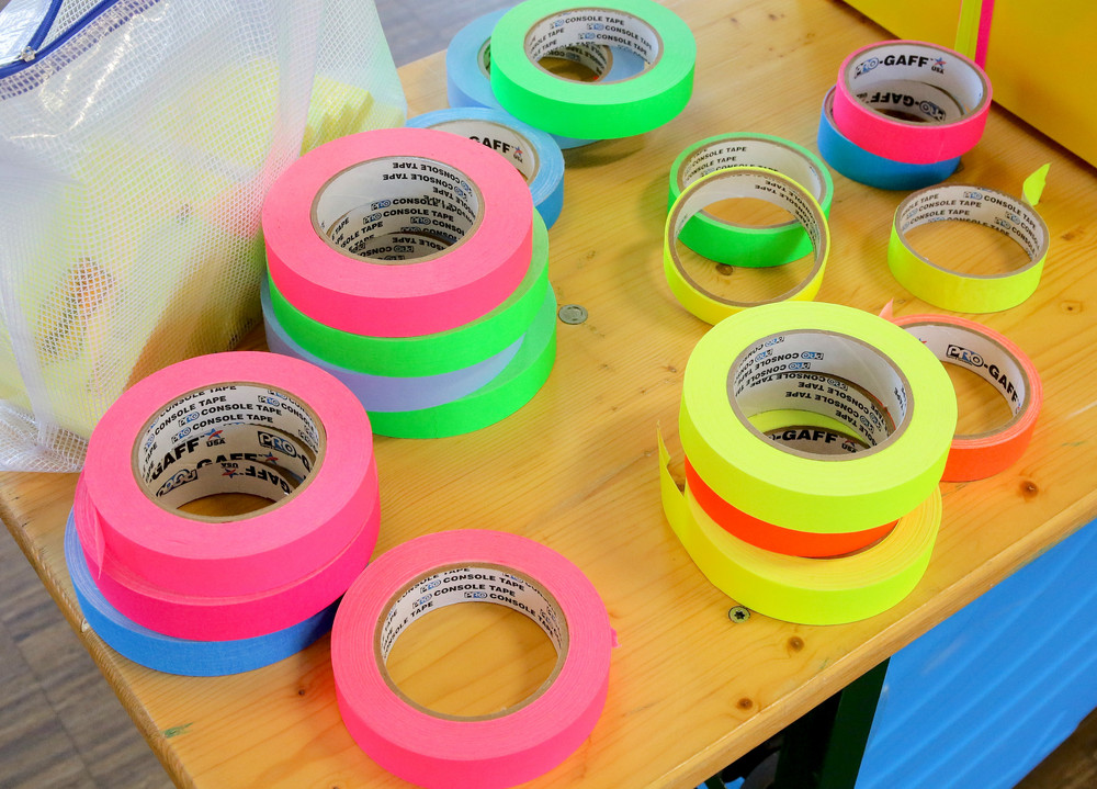 Mehrere Rollen neonfarbener Klebebänder in verschiedenen Farben und Größen liegen auf einem Holztisch neben einem weißen Netzbeutel.