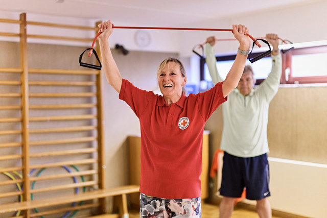 Zwei Personen in einem Sportraum halten ein rotes Fitnessband mit schwarzen Griffen über dem Kopf