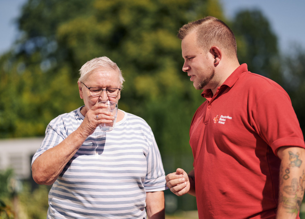 Mitarbeiter des DRK reicht einer Seniorin ein Glas Wasser.