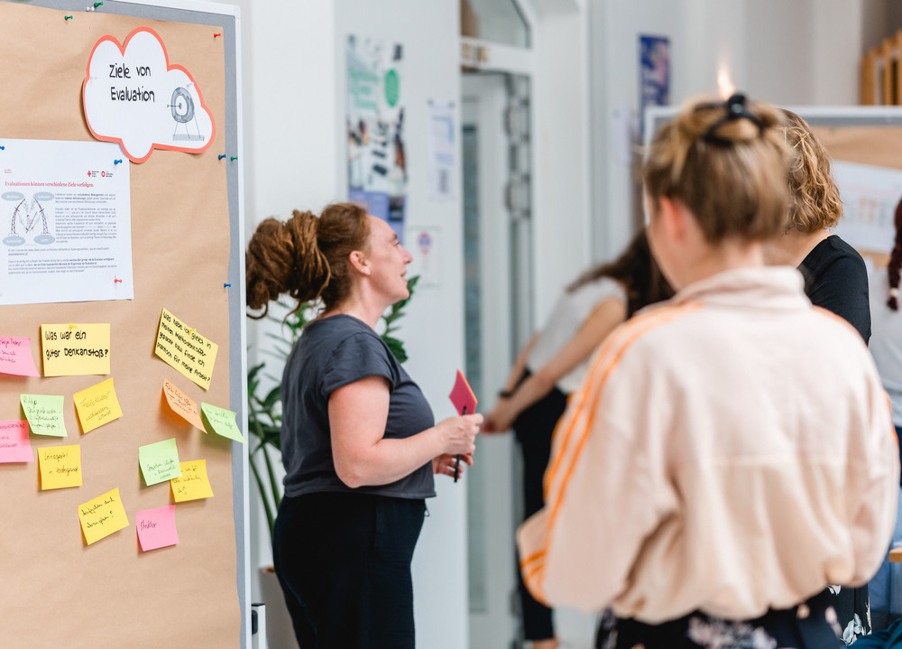 Drei Frauen stehen vor einer Pinnwand auf der die Ziele von Evaluation erklärt werden.