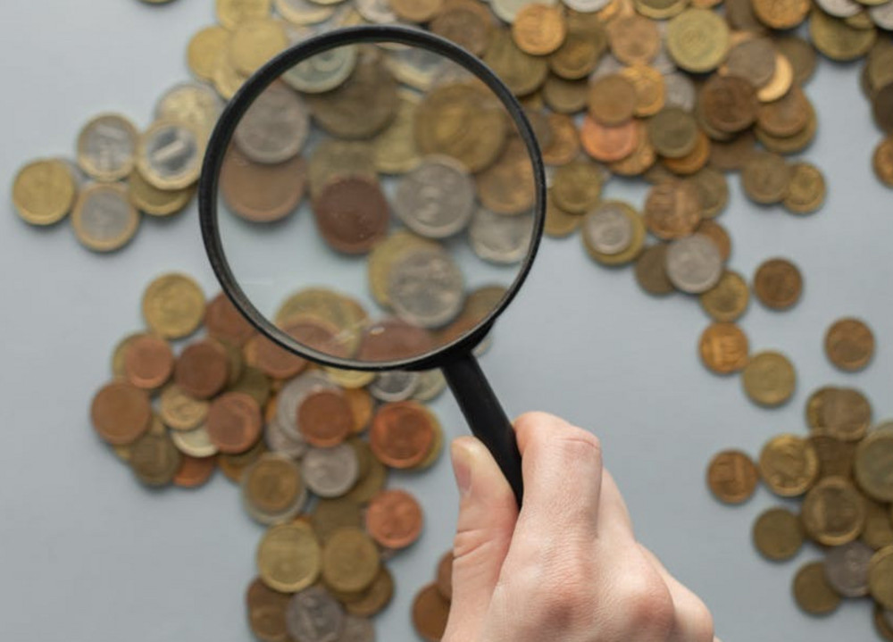 Das Bild zeigt einen auf einem Tisch ausgebreiten Haufen Münzen, die durch eine Lupe betrachtet und näher fokussiert werden.